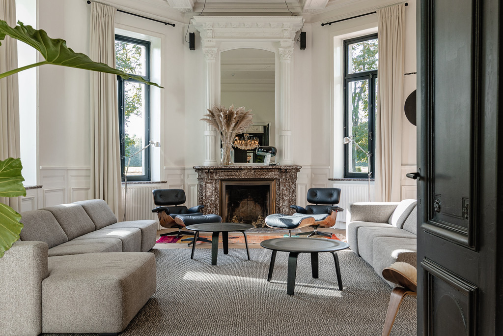 Salon raffiné aux moulures blanches et cheminée en marbre, fauteuils en cuir et canapés gris devant de hautes fenêtres